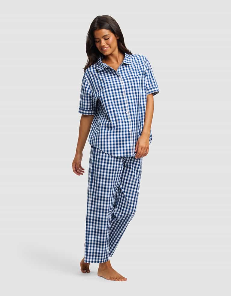 A woman with long dark hair smiles while looking down, barefoot in the Cozy Earth Women's Sutton Short Sleeve & Pant Pajama Set—a blue and white checkered collared button-up shirt and loose pants.|Color:Pacific Gingham