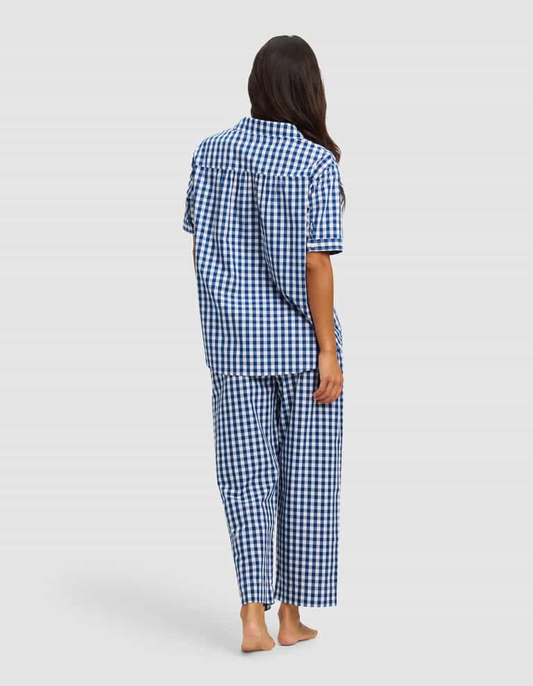 A woman with long brown hair stands barefoot, facing away, wearing Cozy Earth’s Women's Sutton Short Sleeve & Pant Pajama Set in blue and white checks against a plain light background.|Color:Pacific Gingham