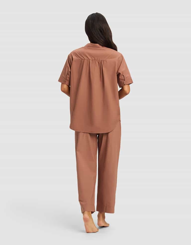 A woman with long dark hair stands barefoot, facing away, wearing the Cozy Earth Women's Sutton Short Sleeve & Pant Pajama Set in brown against a plain light gray background.|Color:Saddle