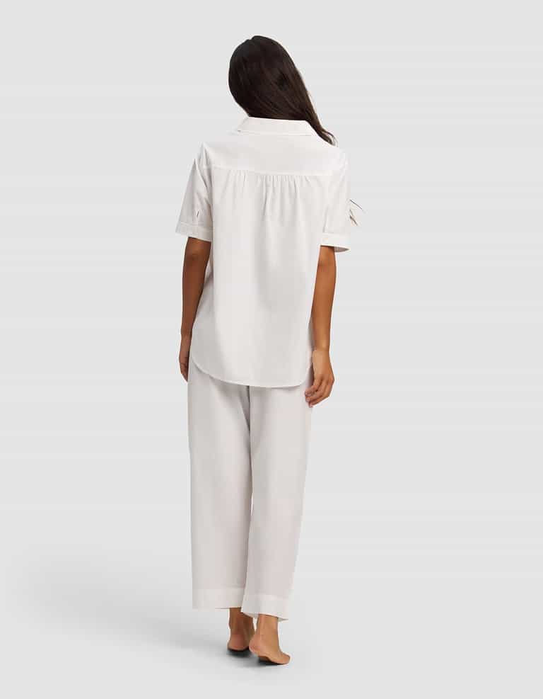 A person with long dark hair stands barefoot, facing away, wearing the Cozy Earth Women's Sutton Short Sleeve & Pant Pajama Set in white against a plain light gray background.|Color:White