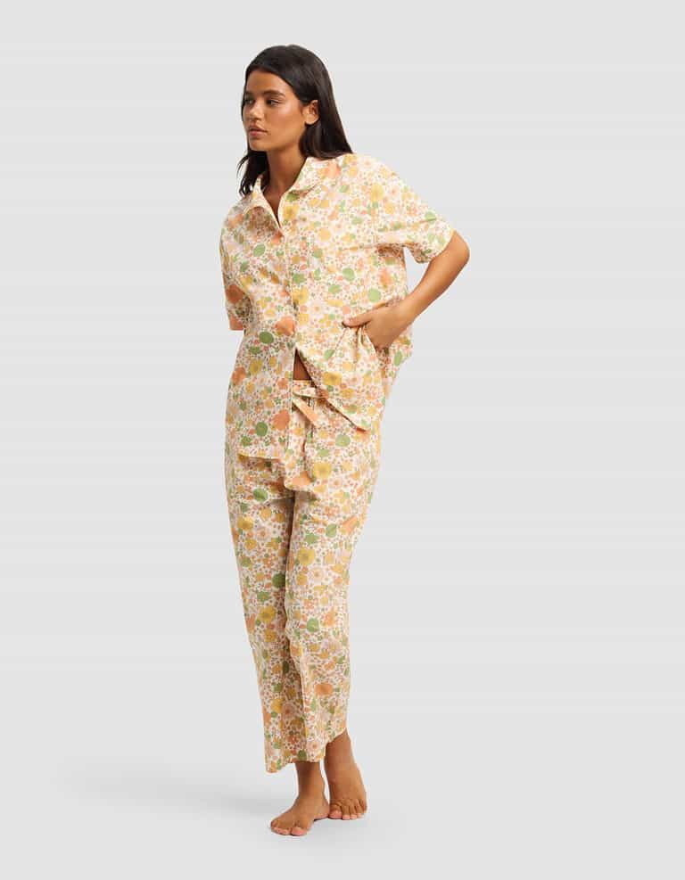 A woman barefoot models the Cozy Earth Women's Sutton Short Sleeve & Pant Pajama Set, featuring a loose-fitting, button-up top and matching pastel floral print pants, with hands in pockets against a plain light gray background.|Color:Wildflower