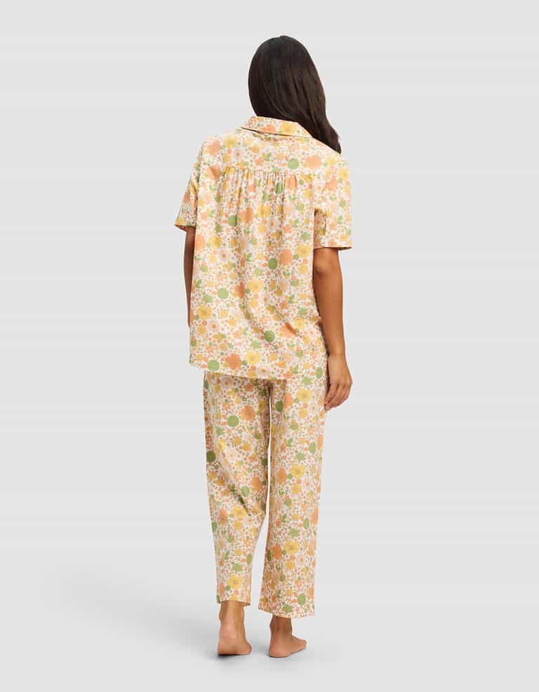 A woman with long brown hair stands barefoot, facing away, wearing Cozy Earth's Women's Sutton Short Sleeve & Pant Pajama Set in a soft orange, green, and white floral pattern on a light background.|Color:Wildflower