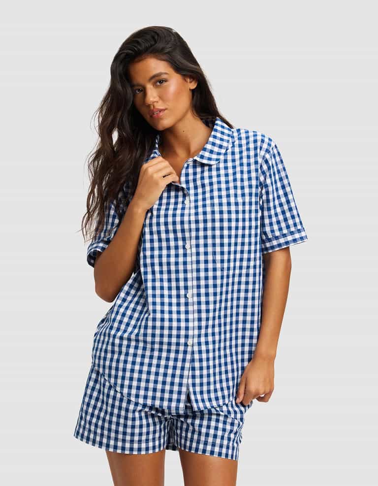A woman with long dark hair wears the Cozy Earth Women's Sutton Short Sleeve Pajama Top, styled in blue and white checks, standing against a light gray background.|Color:Pacific Gingham
