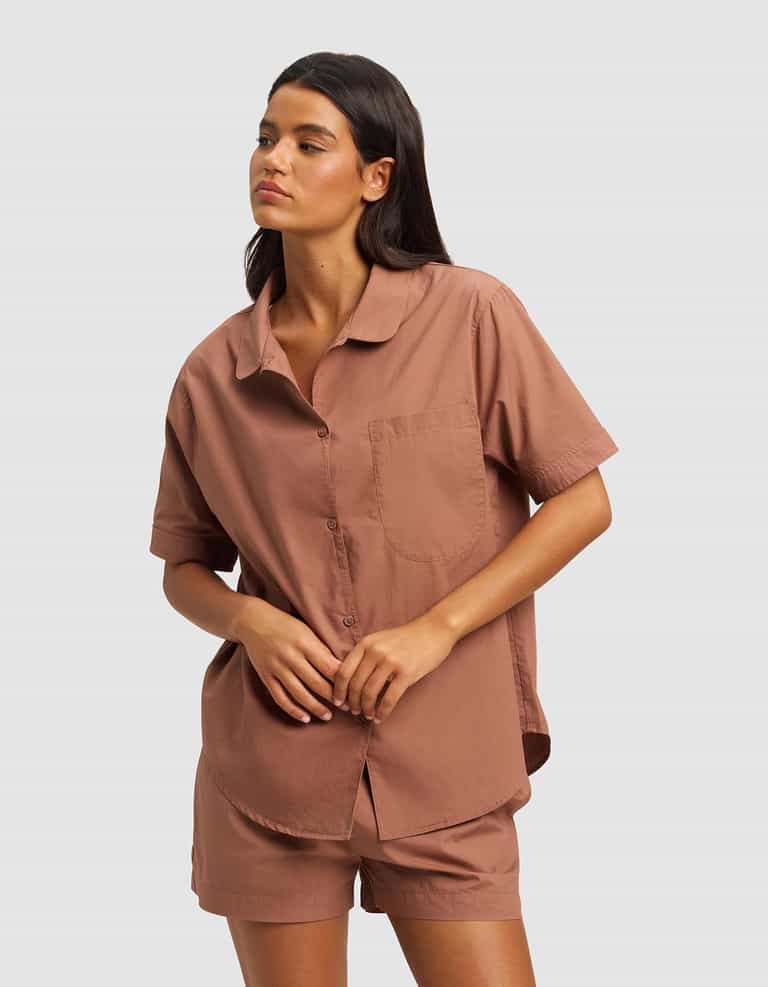 A woman with long dark hair wears the Cozy Earth Women's Sutton Short Sleeve Pajama Top in brown and matching shorts, standing against a plain light gray background.|Color:Saddle