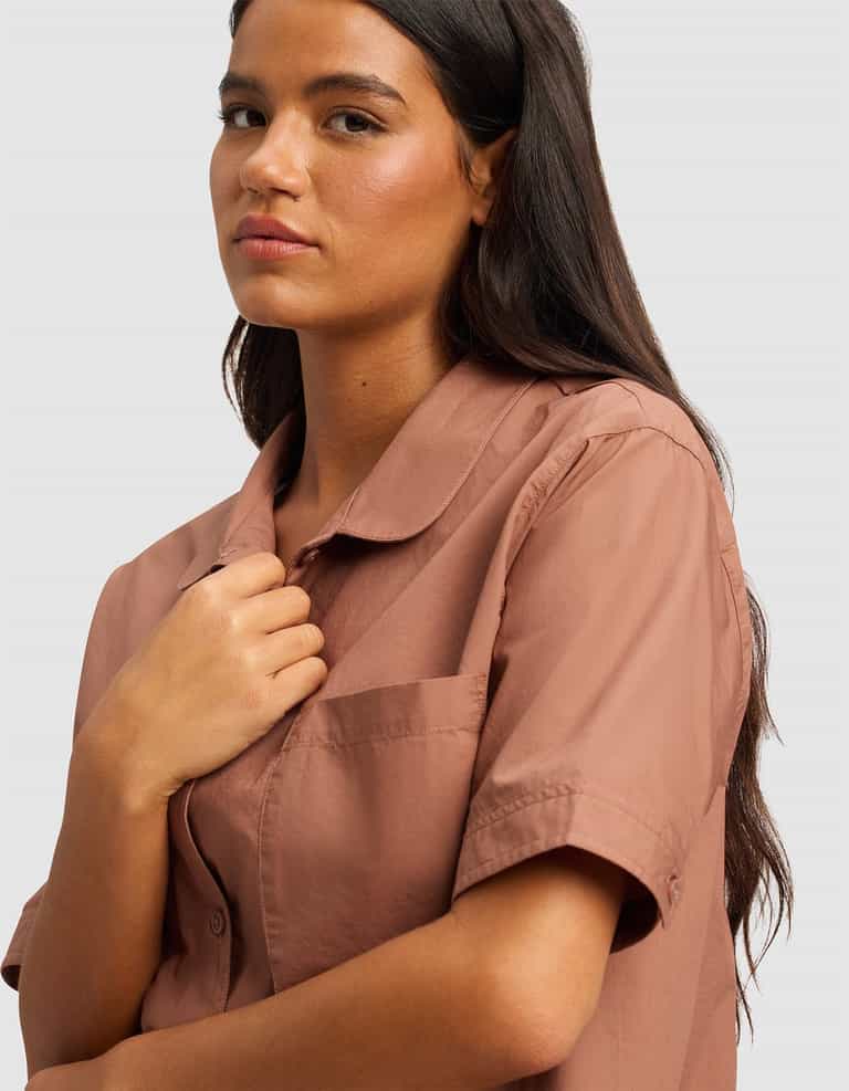 A woman with long dark hair stands against a light gray background, slightly looking to the side, wearing the Cozy Earth Women's Sutton Short Sleeve Pajama Top in light brown and touching the collar.|Color:Saddle