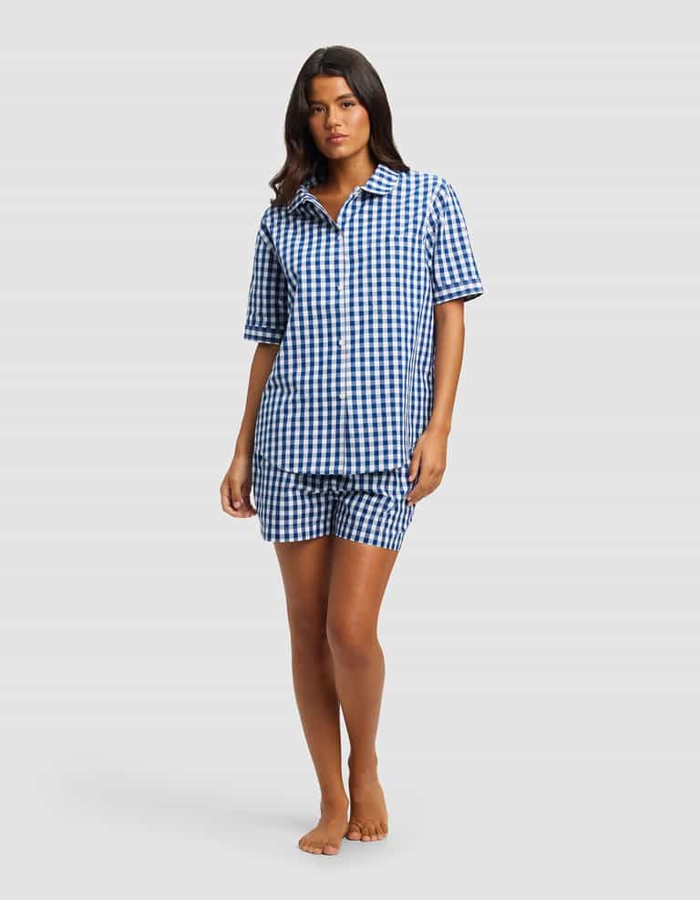 A woman stands barefoot against a plain light gray background wearing the Cozy Earth Women's Sutton Short Sleeve Pajama Set, featuring a matching blue and white checkered top and shorts.|Color:Pacific Gingham