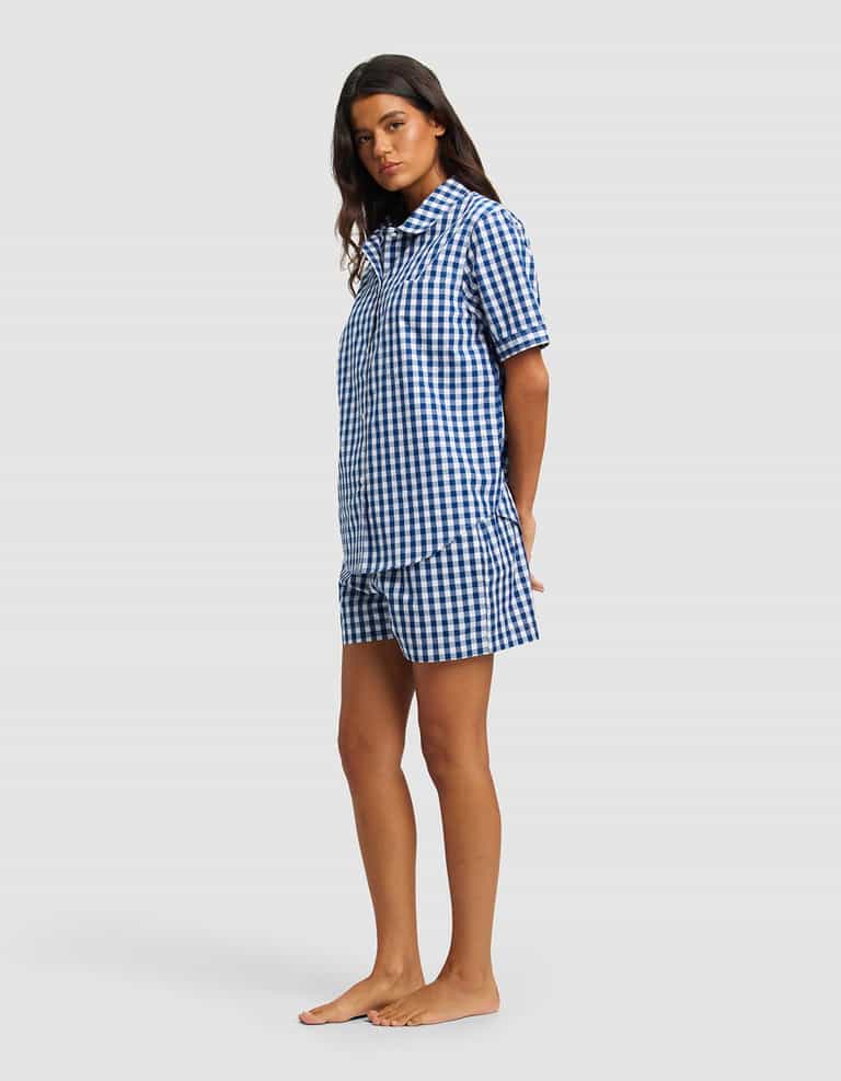 A woman stands barefoot against a plain light background, wearing the Cozy Earth Women's Sutton Short Sleeve Pajama Set in blue and white gingham, with her arms behind her back and facing slightly to the side.|Color:Pacific Gingham