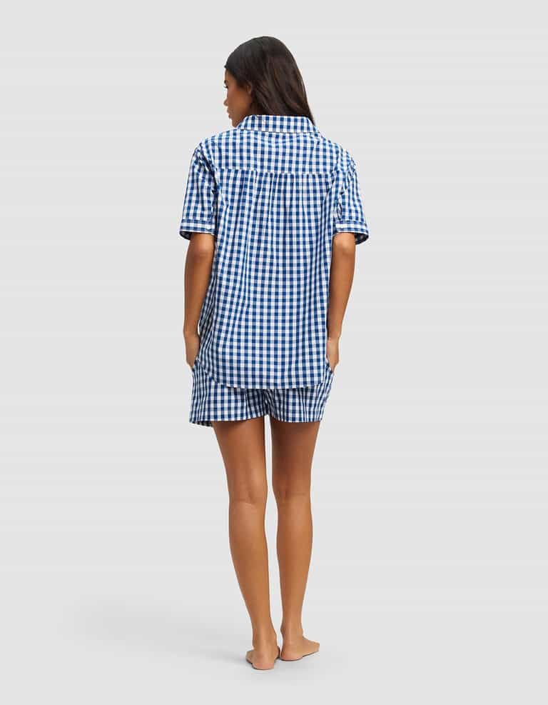 A woman stands barefoot facing away, wearing the Cozy Earth Women's Sutton Short Sleeve Pajama Set in a blue and white checkered pattern. The background is plain light gray.|Color:Pacific Gingham