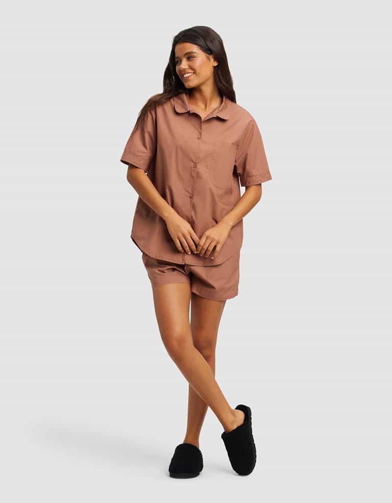 A woman smiles, wearing the Cozy Earth Women's Sutton Short Sleeve Pajama Set in dusty rose, paired with black fuzzy slippers. She poses with one leg crossed over the other against a plain light background.|Color:Saddle