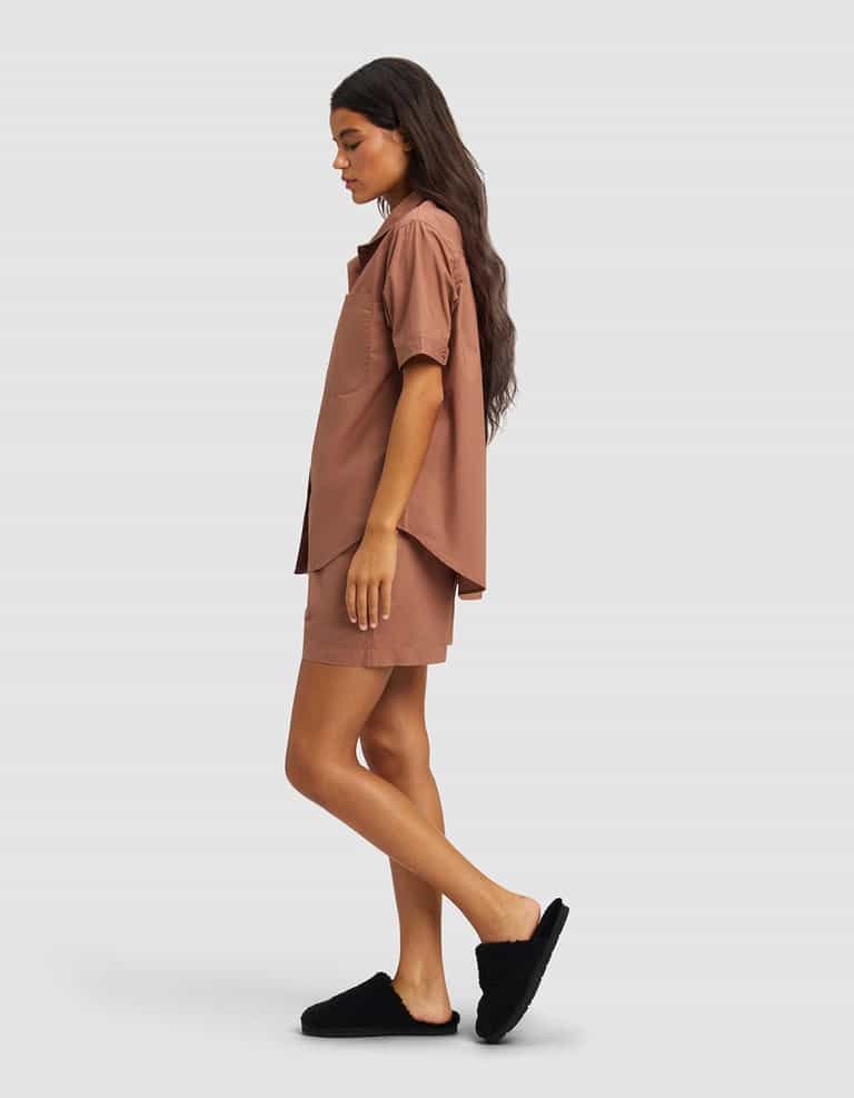 A woman with long dark hair models the Cozy Earth Women's Sutton Short Sleeve Pajama Set in brown, featuring a loose shirt and matching shorts, paired with black slippers, standing in profile against a light gray background.|Color:Saddle