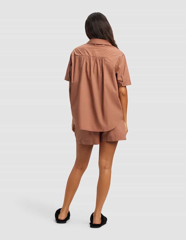 A woman with long brown hair wears the Cozy Earth Women's Sutton Short Sleeve Pajama Set in brown, featuring a relaxed-fit button-up shirt and matching shorts. She stands facing away in black slip-on shoes against a plain light gray background.|Color:Saddle