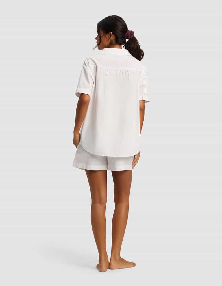 A woman with dark hair in a scrunchie stands barefoot, facing away, wearing the Cozy Earth Women's Sutton Short Sleeve Pajama Set—loose white shirt and matching shorts—against a plain light background.|Color:White