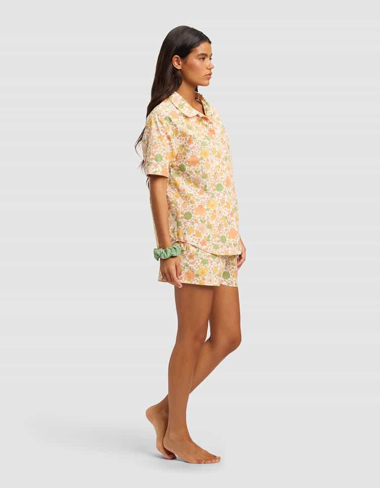 A woman with long brown hair stands barefoot in profile, wearing the Cozy Earth Women's Sutton Short Sleeve Pajama Set in pastel floral, with a light green scrunchie on her wrist, posing against a plain gray background.|Color:Wildflower