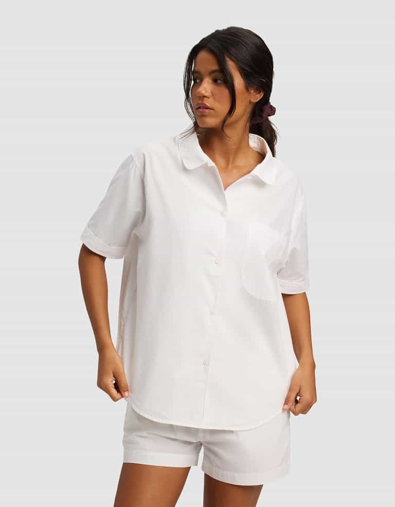 A woman wears the Cozy Earth Women's Sutton Short Sleeve Pajama Top, a loose-fitting white button-up shirt with a chest pocket, paired with matching white shorts, standing against a plain light gray background.|Color:White