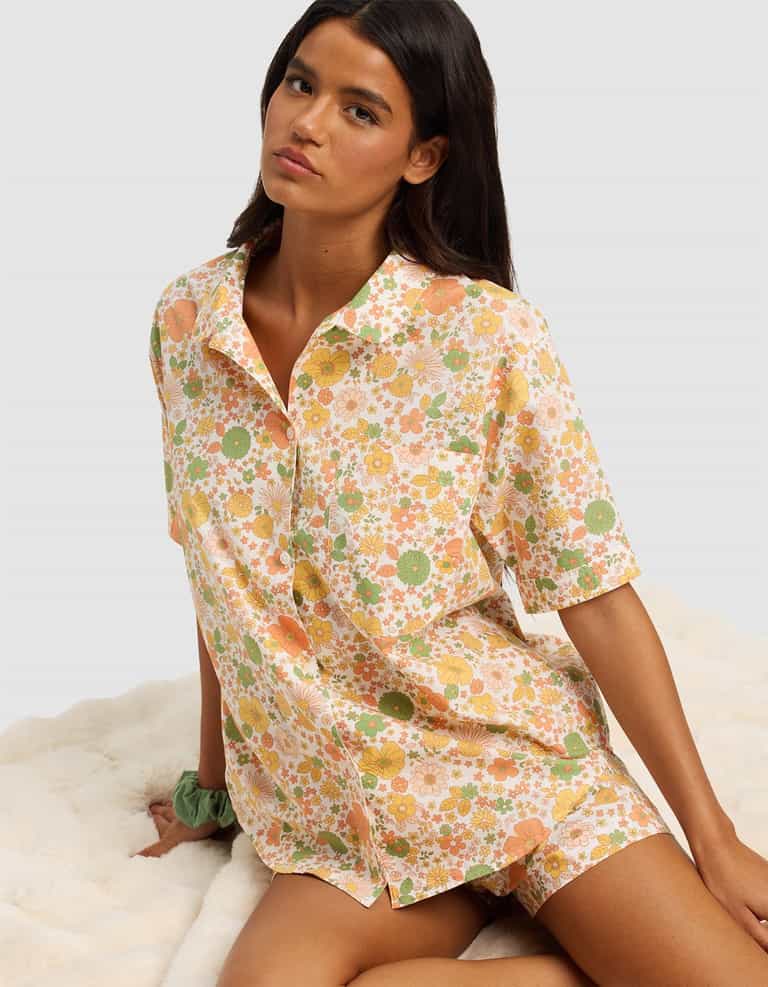 A woman with long dark hair sits on a white fuzzy surface, wearing the Cozy Earth Women's Sutton Short Sleeve Pajama Top featuring a colorful floral pattern in orange, yellow, and green.|Color:Wildflower