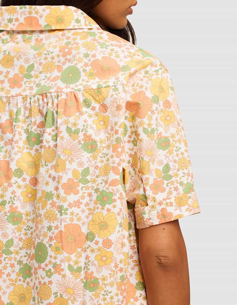 A woman is shown from the back wearing the Cozy Earth Women's Sutton Short Sleeve Pajama Top, featuring a pastel floral pattern in yellow, green, and peach against a plain light background.|Color:Wildflower