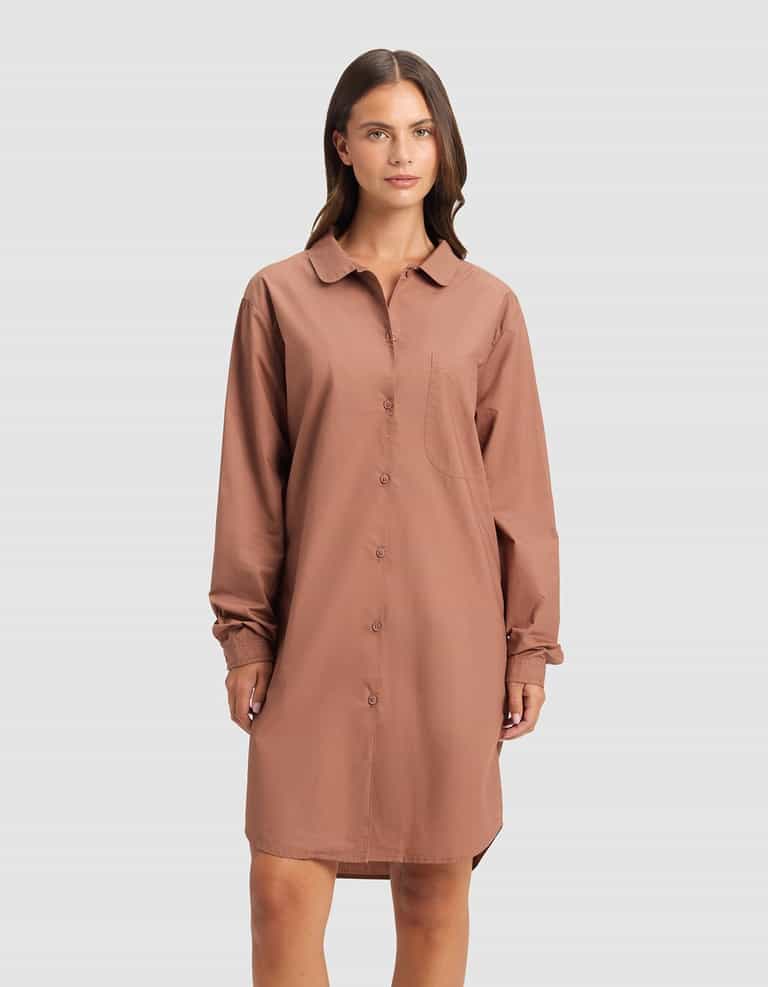 A woman stands against a plain background wearing the Cozy Earth Women's Sutton Sleep Dress, a loose, long-sleeve, button-down brown shirt dress with a collar and chest pocket.|Color:Saddle