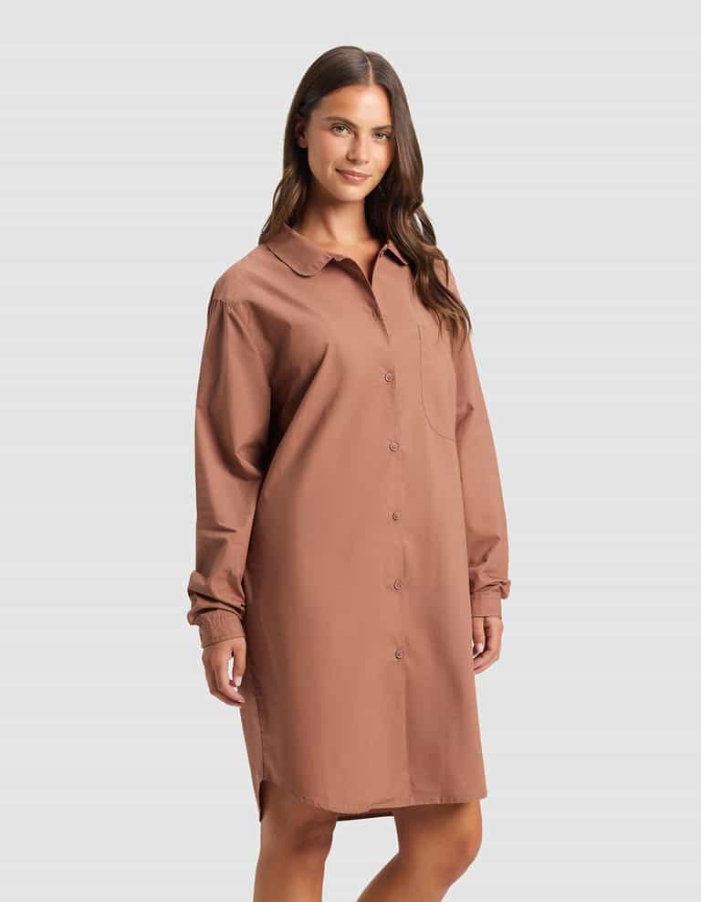 A woman smiles slightly against a plain light gray background, wearing the Cozy Earth Women's Sutton Sleep Dress—a loose-fitting, long-sleeve, button-down shirt dress in brownish-pink with a collared neckline and chest pocket.|Color:Saddle