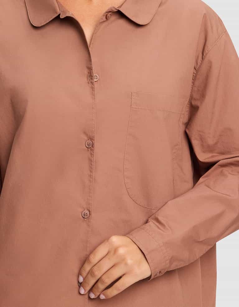 A person shown from shoulders to hips wears the Cozy Earth Women's Sutton Sleep Dress, a loose, long-sleeve, button-up sleep shirt in muted brown with a front pocket. Their hand rests near the buttons.|Color:Saddle
