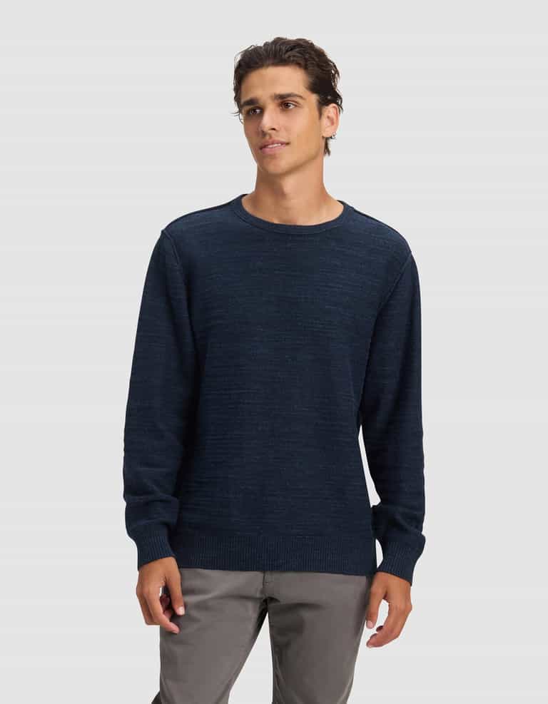A young man with short, wavy brown hair wears the Cozy Earth Men's Tate Crewneck in dark blue and light gray pants, standing against a plain light gray background with a neutral expression, gazing slightly to the side.|Color:Eclipse