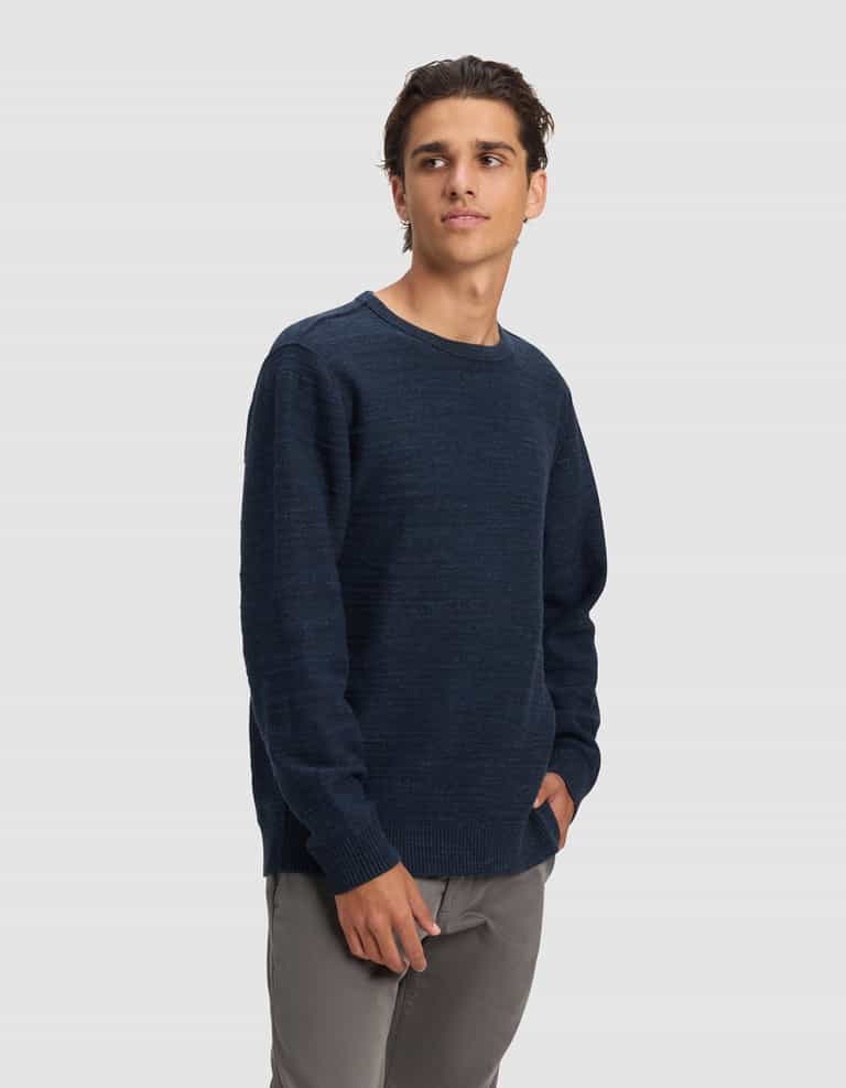 A young man with dark hair wears the Cozy Earth Men's Tate Crewneck in navy blue and gray pants, standing against a plain light gray background, gazing slightly left with his right hand in his pocket.|Color:Eclipse