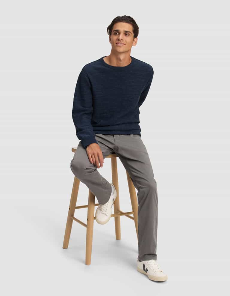 A young man with short, dark hair sits on a wooden stool against a plain background, relaxed and wearing the Cozy Earth Men's Tate Crewneck in navy blue, light gray pants, and white sneakers.|Color:Eclipse