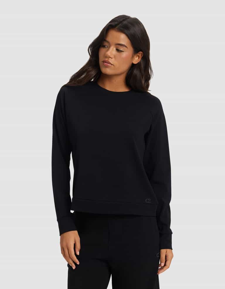 A woman with long dark hair wears the Cozy Earth Women's Brushed Bamboo Pullover Crew and matching black pants, standing against a light gray background and looking slightly to her left.|Color:Black