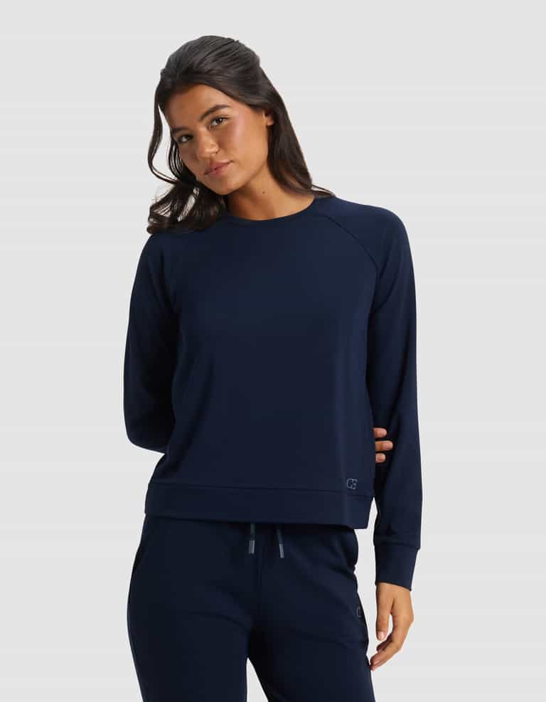 A woman with long dark hair wears the Cozy Earth Women's Brushed Bamboo Pullover Crew in navy blue, paired with matching pants, standing against a plain light gray background. She looks slightly to the side with a relaxed expression.|Color:Navy
