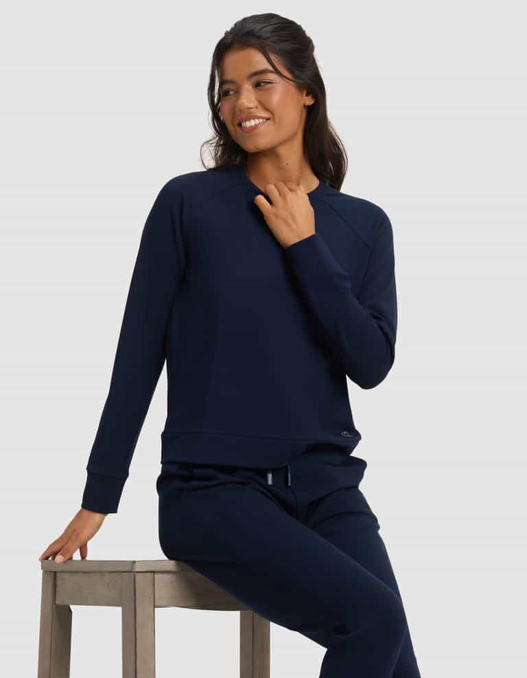 A woman in a Cozy Earth Women's Brushed Bamboo Pullover Crew sits on a wooden stool, smiling and looking to the side against a plain light background.|Color:Navy