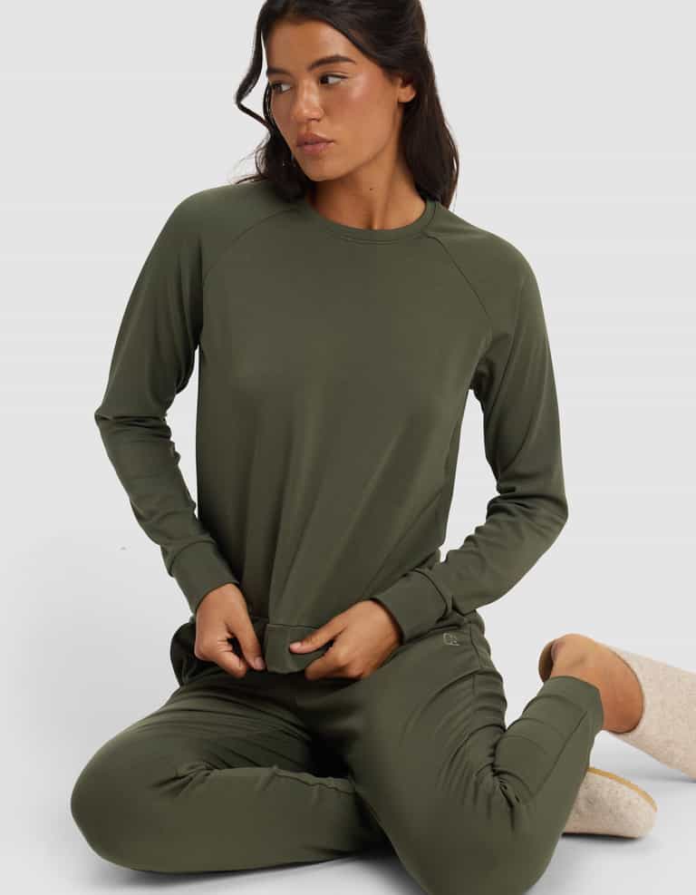 A woman sits on the floor with one knee bent, looking to the side, wearing Cozy Earth’s Women’s Brushed Bamboo Pullover Crew in olive green against a plain light background.|Color:Olive