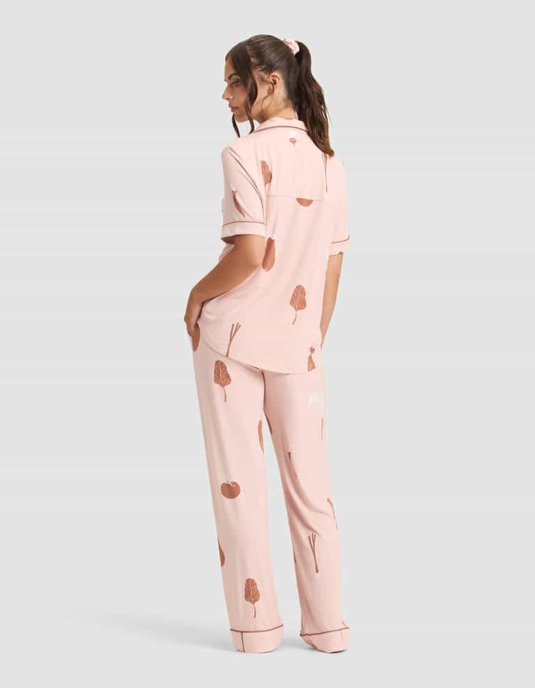 A person faces away in Cozy Earth's Women's Bamboo Stretch-Knit Short Sleeve & Pant Pajama Set—light pink with a brown tree and apple print, featuring a collared short-sleeve top and loose long pants, against a plain gray background. |Color:Veggies in Dusty Rose