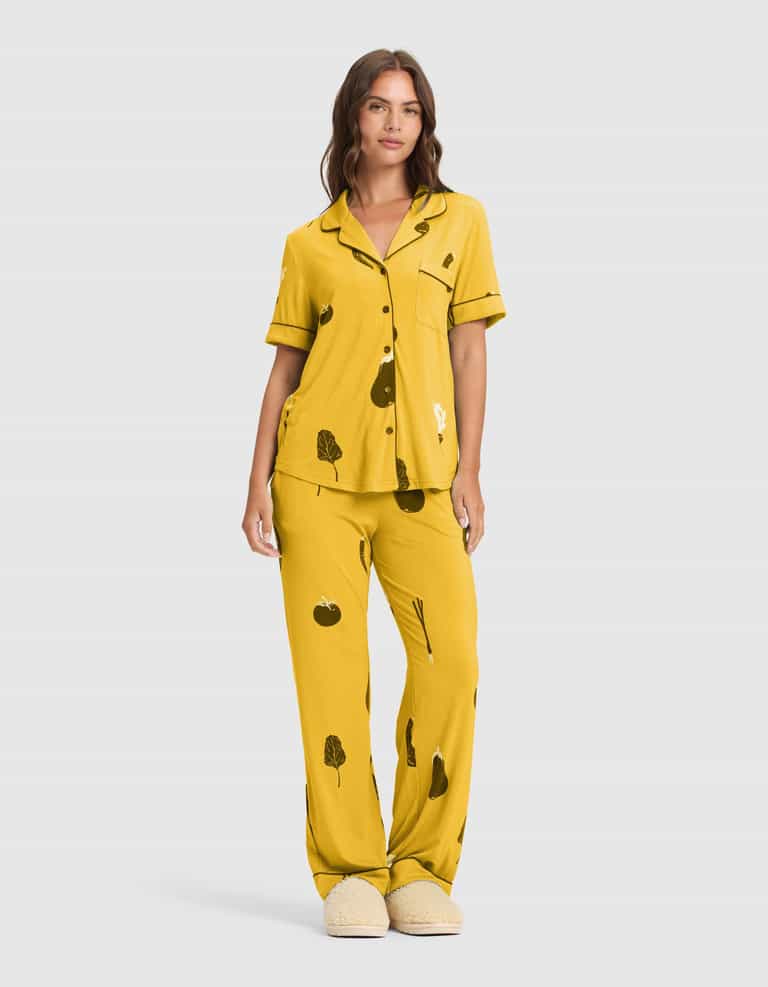 A woman wears the Cozy Earth Women's Bamboo Stretch-Knit Short Sleeve & Pant Pajama Set (Veggie) featuring a yellow veggie print, button-front top, and matching slippers. She has long brown hair and a neutral expression. |Color:Veggies in Mango