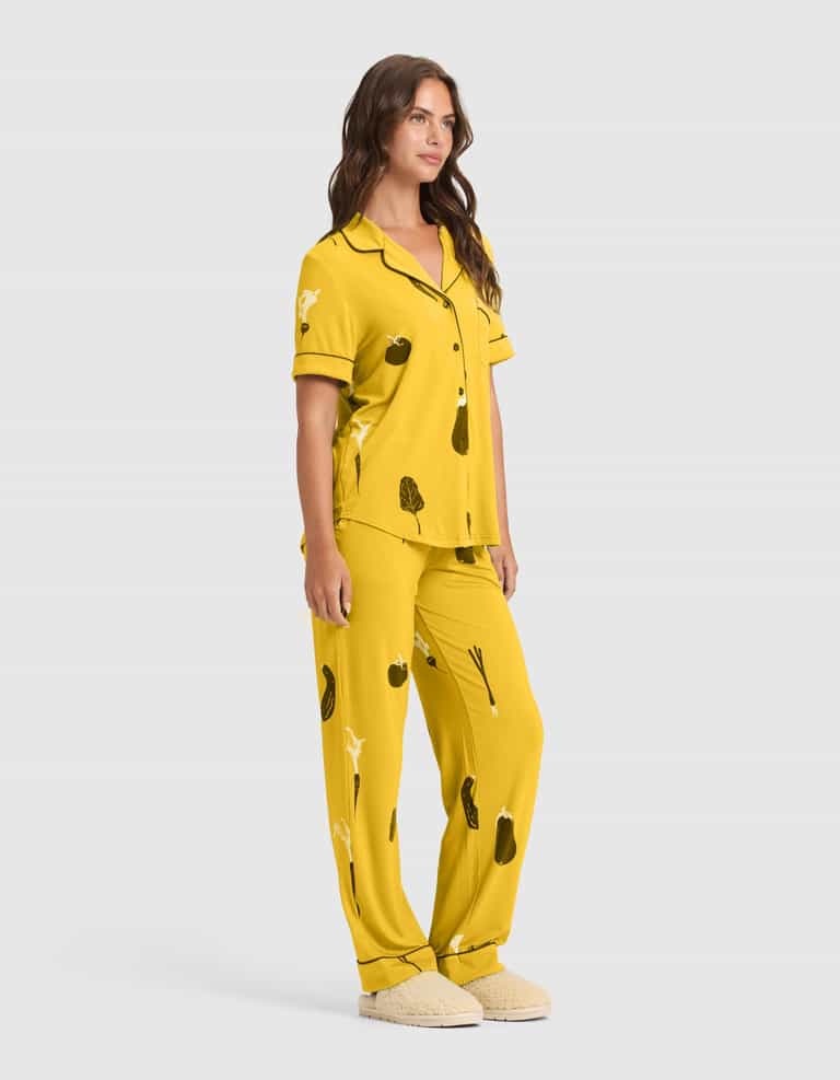 A woman models the Cozy Earth Women's Bamboo Stretch-Knit Short Sleeve & Pant Pajama Set (Veggie), featuring black vegetable prints and matching slippers, posing with a relaxed expression against a plain light background. |Color:Veggies in Mango