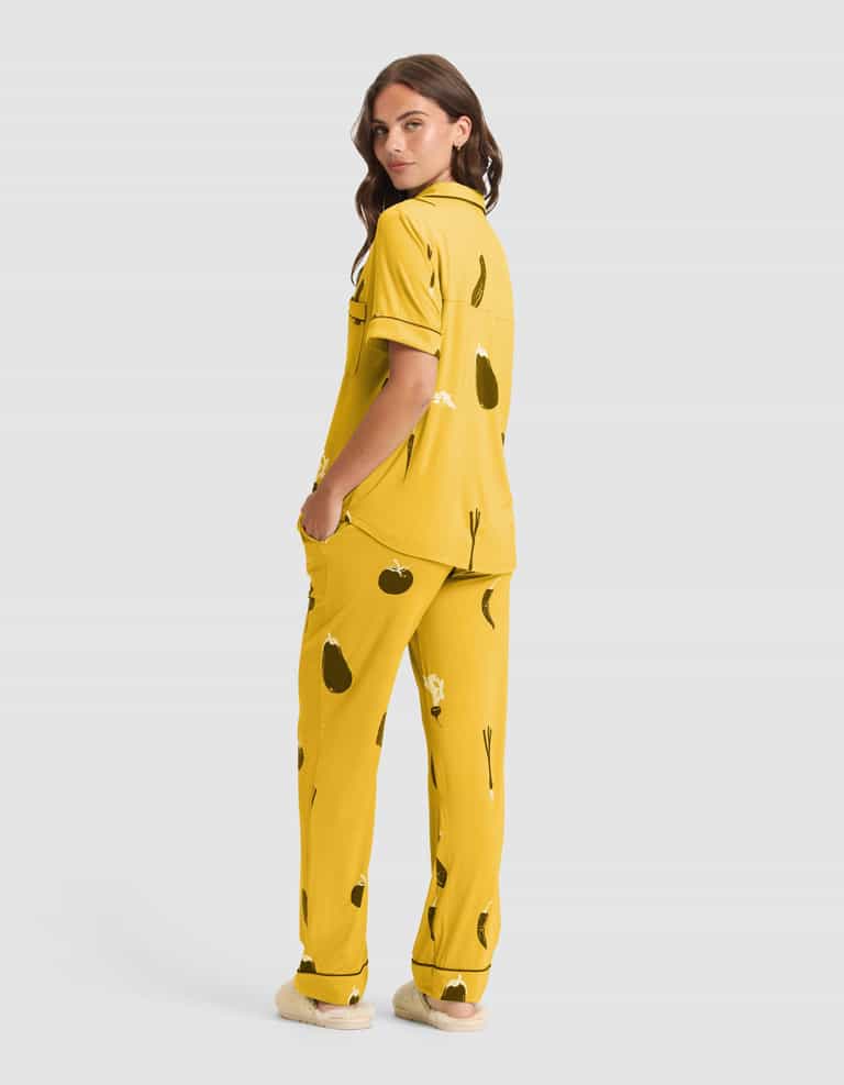 A woman faces backward, head turned to the camera, modeling Cozy Earth's Women's Bamboo Stretch-Knit Short Sleeve & Pant Pajama Set (Veggie) with matching slippers against a plain light gray background. |Color:Veggies in Mango