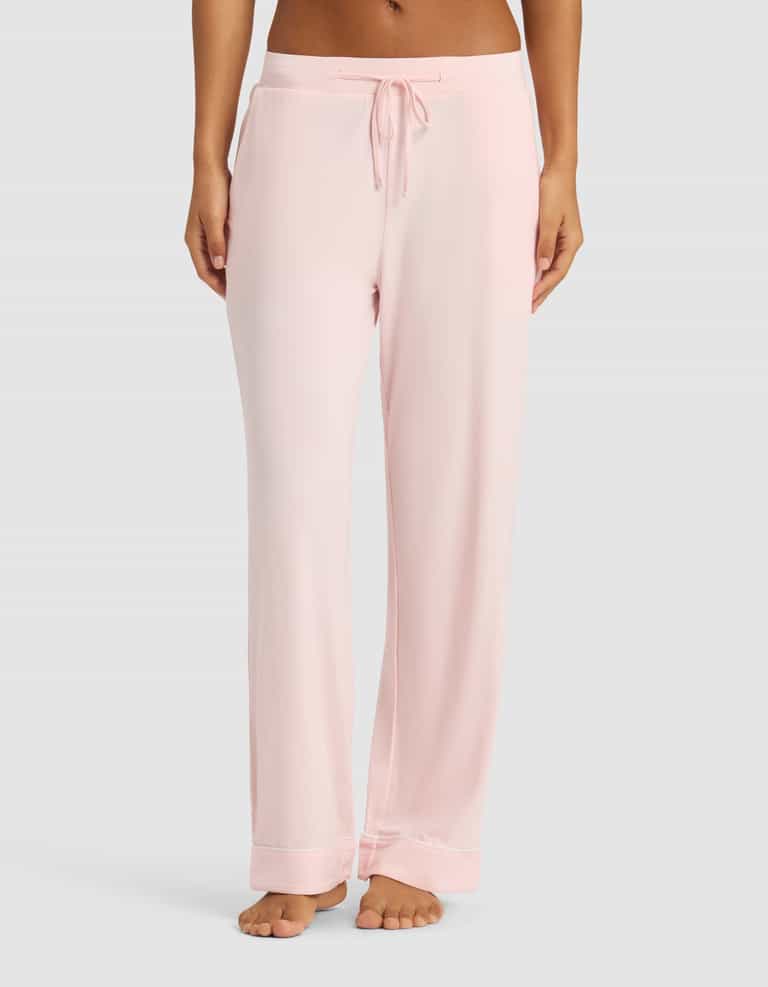 A woman wears Cozy Earth's Women's Bamboo Stretch-Knit Long Sleeve Pajama Set in light pink with a drawstring waist, standing barefoot against a plain light gray background.|Color:Blossom