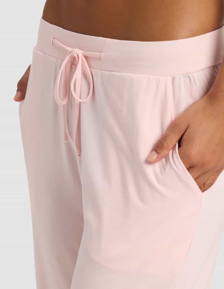A close-up shows a person wearing Cozy Earth Women's Bamboo Stretch-Knit Pajama Pants in light pink, featuring a drawstring tied at the waist and one hand in the pocket, set against a plain light background.|Color:Blossom