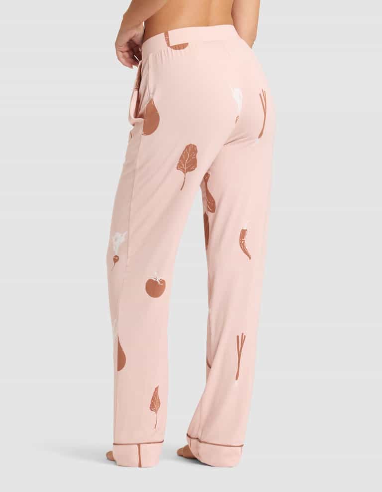 A person is barefoot wearing HIDE Women's Bamboo Stretch-Knit Pajama Pant in light pink with brown and white vegetable prints, shown from the lower back and legs against a plain light gray background. |Color:Veggies in Dusty Rose
