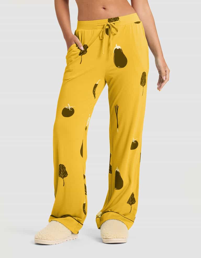 A woman wears Cozy Earth's Women's Bamboo Stretch-Knit Short Sleeve & Pant Pajama Set (Veggie), featuring loose-fitting yellow veggie-print pants with pockets and a drawstring waist, paired with cream slippers against a plain light gray background. |Color:Veggies in Mango