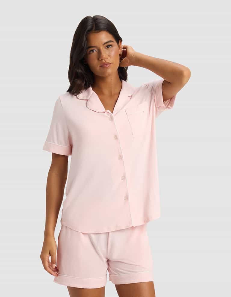 A woman wears the Cozy Earth Women's Bamboo Stretch-Knit Short Sleeve Pajama Top in light pink with matching shorts, standing against a plain light gray background, one hand touching her hair.|Color:Blossom