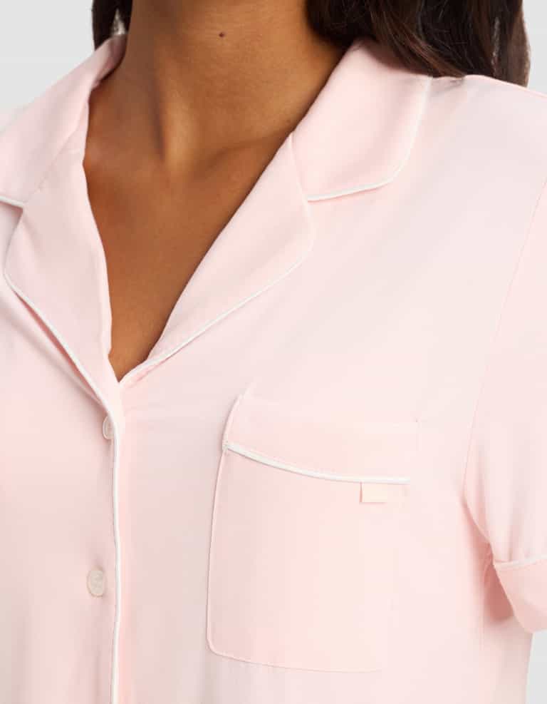 Close-up of a person wearing the Cozy Earth Women's Bamboo Stretch-Knit Short Sleeve Pajama Top in light pink with white piping, chest pocket, and partially visible buttons. The person's face is not shown.|Color:Blossom