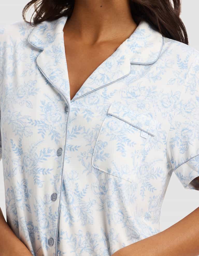 A woman wears the Cozy Earth Women's Bamboo Stretch-Knit Short Sleeve Pajama Top, featuring white fabric with light blue floral prints, grey piping, buttons, a chest pocket, and a notched collar. Shown from shoulders to waist.|Color:Sky Toile