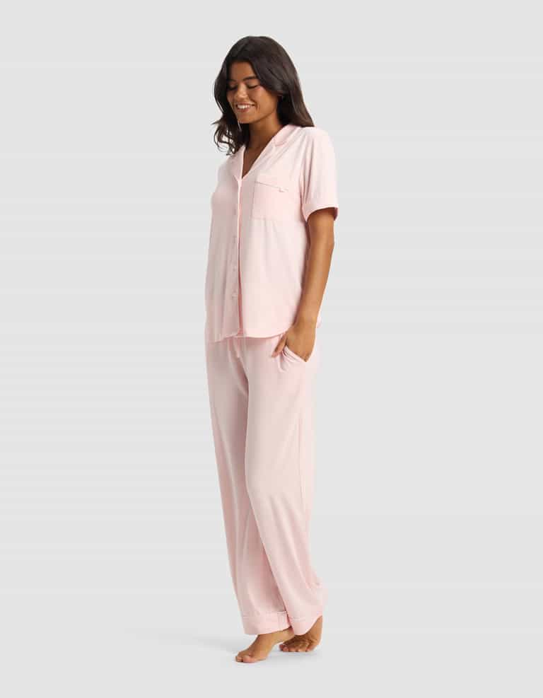 A woman stands barefoot, smiling, in the Cozy Earth Women's Bamboo Stretch-Knit Short Sleeve & Pant Pajama Set in light pink, with her left hand in her pocket against a plain light gray background.|Color:Blossom