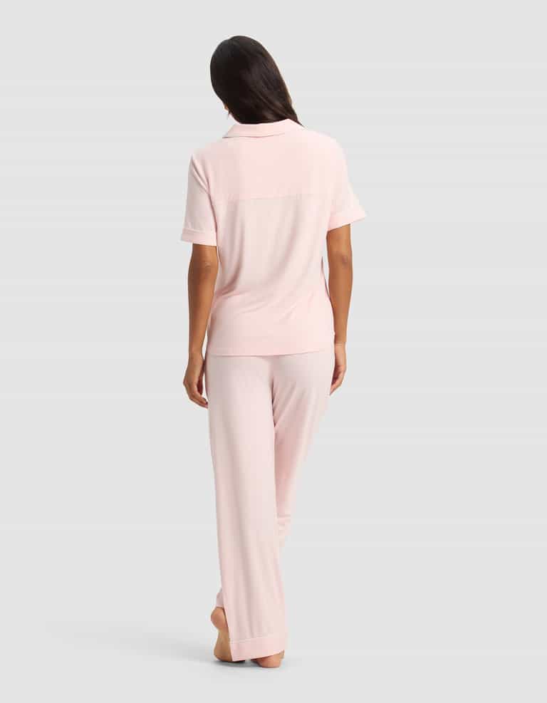 A woman stands barefoot, facing away, wearing the Cozy Earth Women's Bamboo Stretch-Knit Short Sleeve & Pant Pajama Set in light pink, against a plain gray background.|Color:Blossom