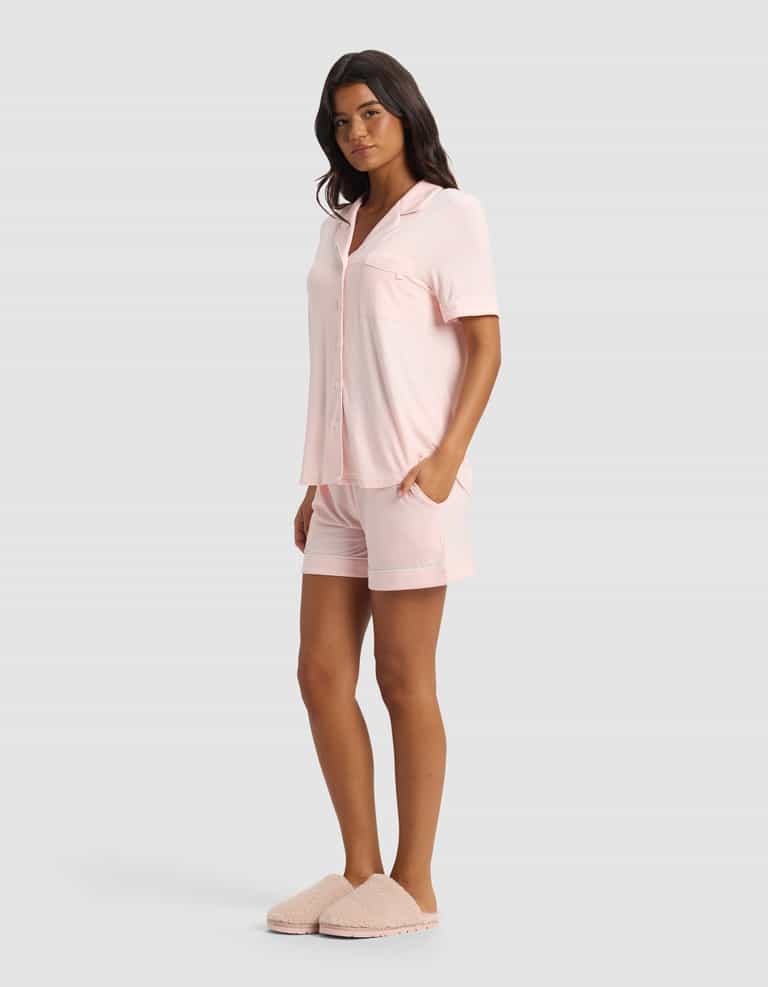 A woman wears the Cozy Earth Women's Bamboo Stretch-Knit Short Sleeve Pajama Set in pale pink with matching shorts and fluffy slippers, posing against a plain light gray background.|Color:Blossom