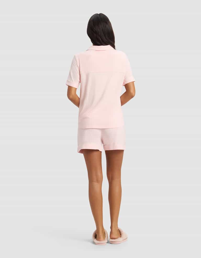 A woman faces away, wearing Cozy Earth’s Women's Bamboo Stretch-Knit Short Sleeve Pajama Set in light pink with matching shorts and pink slippers, set against a plain light gray background.|Color:Blossom