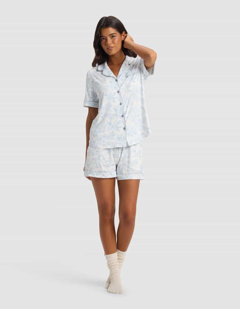 A woman stands in a relaxed pose wearing the Cozy Earth Women's Bamboo Stretch-Knit Short Sleeve Pajama Set with matching shorts and cream socks against a plain light background.|Color:Sky Toile