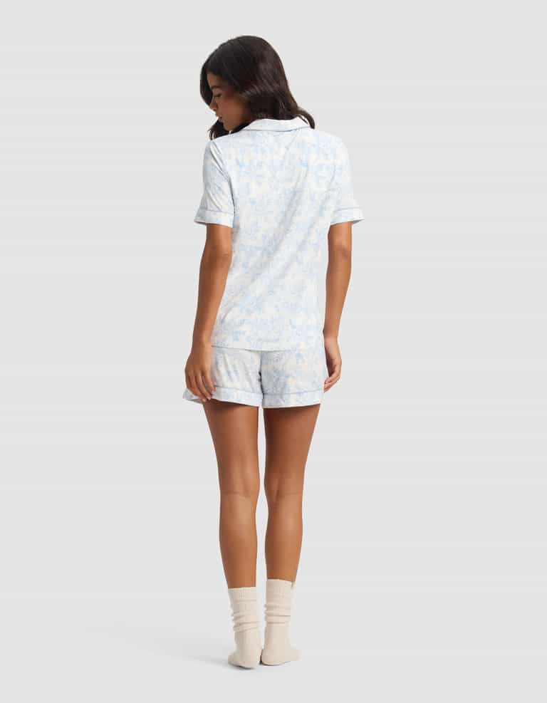 A woman with medium-length dark hair wears the Cozy Earth Women's Bamboo Stretch-Knit Short Sleeve Pajama Set in light blue and white, paired with cream socks, facing away against a plain light background.|Color:Sky Toile