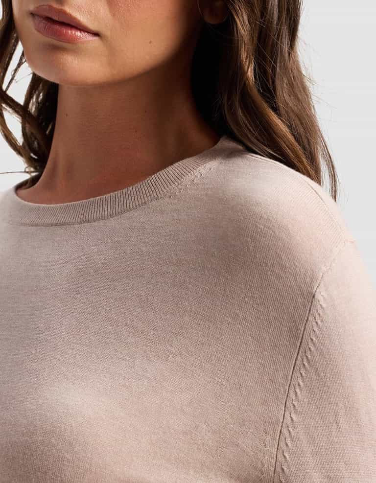 A close-up of a woman wearing the Cozy Earth Women's AirKnit Crewneck Sweater in light beige, showing her shoulder, part of her face, and wavy brown hair against a plain background.|Color:Taupe