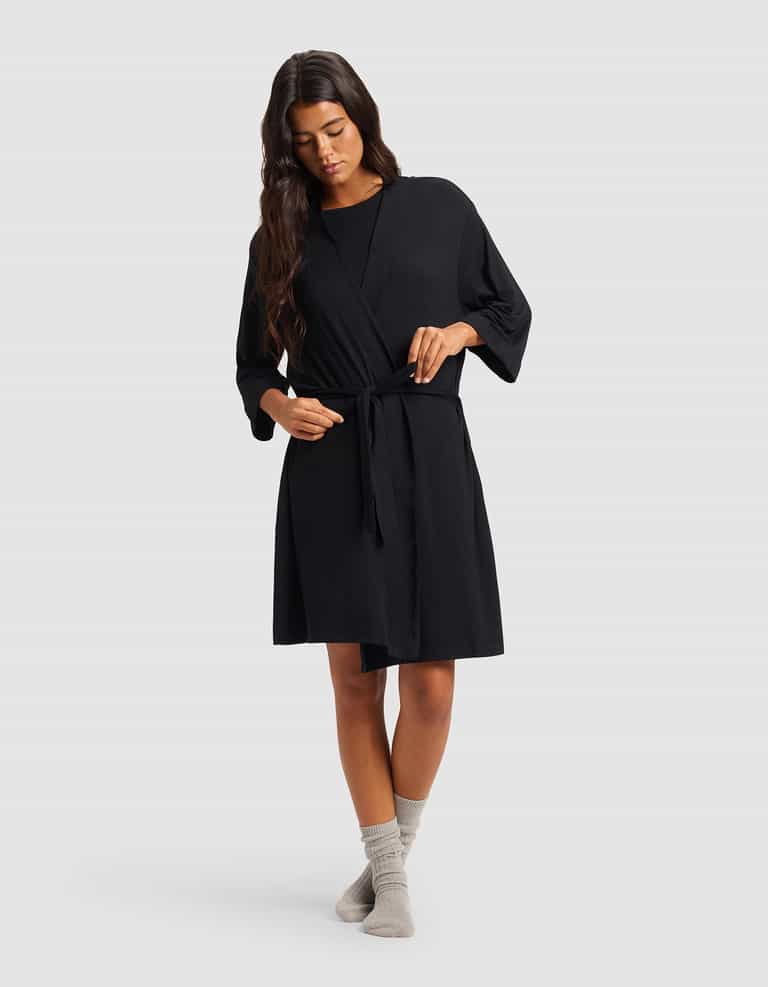 A woman with long dark hair wears the Cozy Earth Women's Bamboo Stretch-Knit Kimono Robe in black, tied at the waist, along with light gray socks. She stands against a plain light gray background, looking down as she gently adjusts the robe's belt.|Color:Black