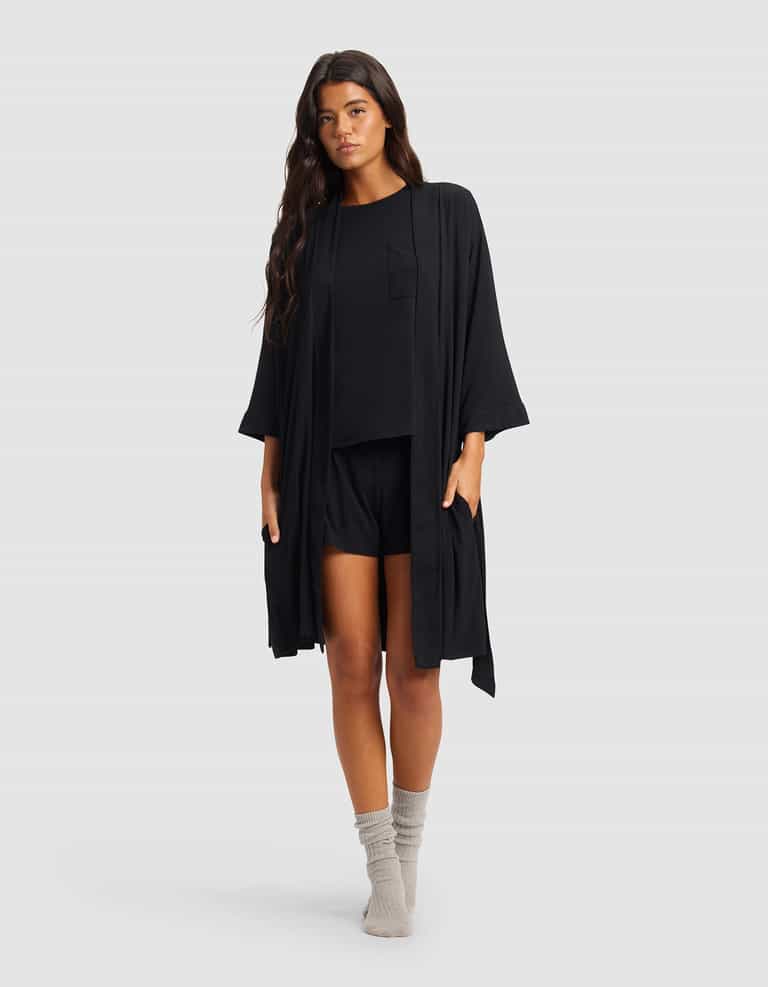 A woman with long dark hair wears the Cozy Earth Women's Bamboo Stretch-Knit Kimono Robe over a black top and shorts, paired with light gray socks, standing against a plain light gray background.|Color:Black